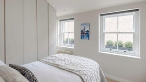 A bedroom with a bed, a radiator, and two windows with a cityscape painting. - Stunning 2-Bed Mews House w/ AC near Regent's Park (London)