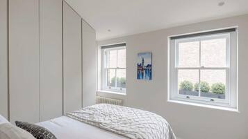 A bedroom with a bed, a radiator, and two windows with a cityscape painting.