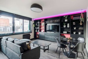 A modern living room with a sofa, coffee table, and chairs, a TV, and shelves with plants and books.