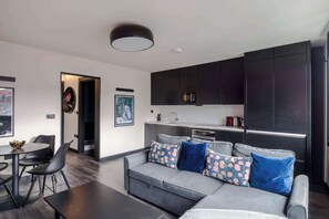 A modern living room with a grey sofa, black chairs, a dining table, and a kitchen area with dark cabinets and a microwave.