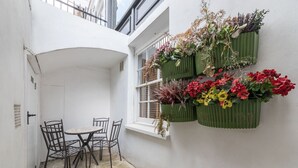 A small patio with a table and chairs, a window with a view, and green planters with flowers. - Fitzrovia Duplex · 2-Bed · Private Patio Garden (London)