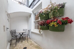 Eine kleine Terrasse mit Tisch und Stühlen, ein Fenster mit Aussicht und grüne Pflanzgefäße mit Blumen.