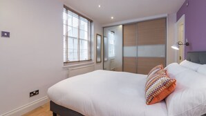 A bedroom with a bed, a window with blinds, a mirror, a wardrobe, and a bedside lamp. - Fitzrovia Duplex · 2-Bed · Private Patio Garden (London)