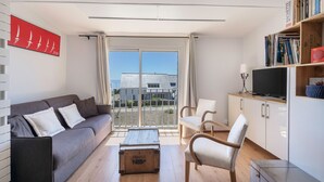 A modern living room with a grey sofa, a wooden coffee table, and a white armchair. There is a bookshelf with books and a television on a stand. A large window with curtains provides a view of buildings outside. - 200 m from the beach, lovely flat for 4 people (Saint-Gildas-de-Rhuys)