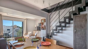 A modern dining area with a wooden table set for four, a staircase with a geometric design, and a view of the outside through large windows. - 200 m from the beach, lovely flat for 4 people (Saint-Gildas-de-Rhuys)