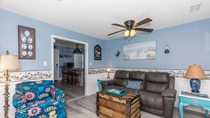 A living room with a blue wall, a brown leather sofa, a floral armchair, a wooden chest, and a ceiling fan. - The Treasure Chest - 3 BR/2 BA Upper Part of Duplex in the Heart of Surf City (Surf City)