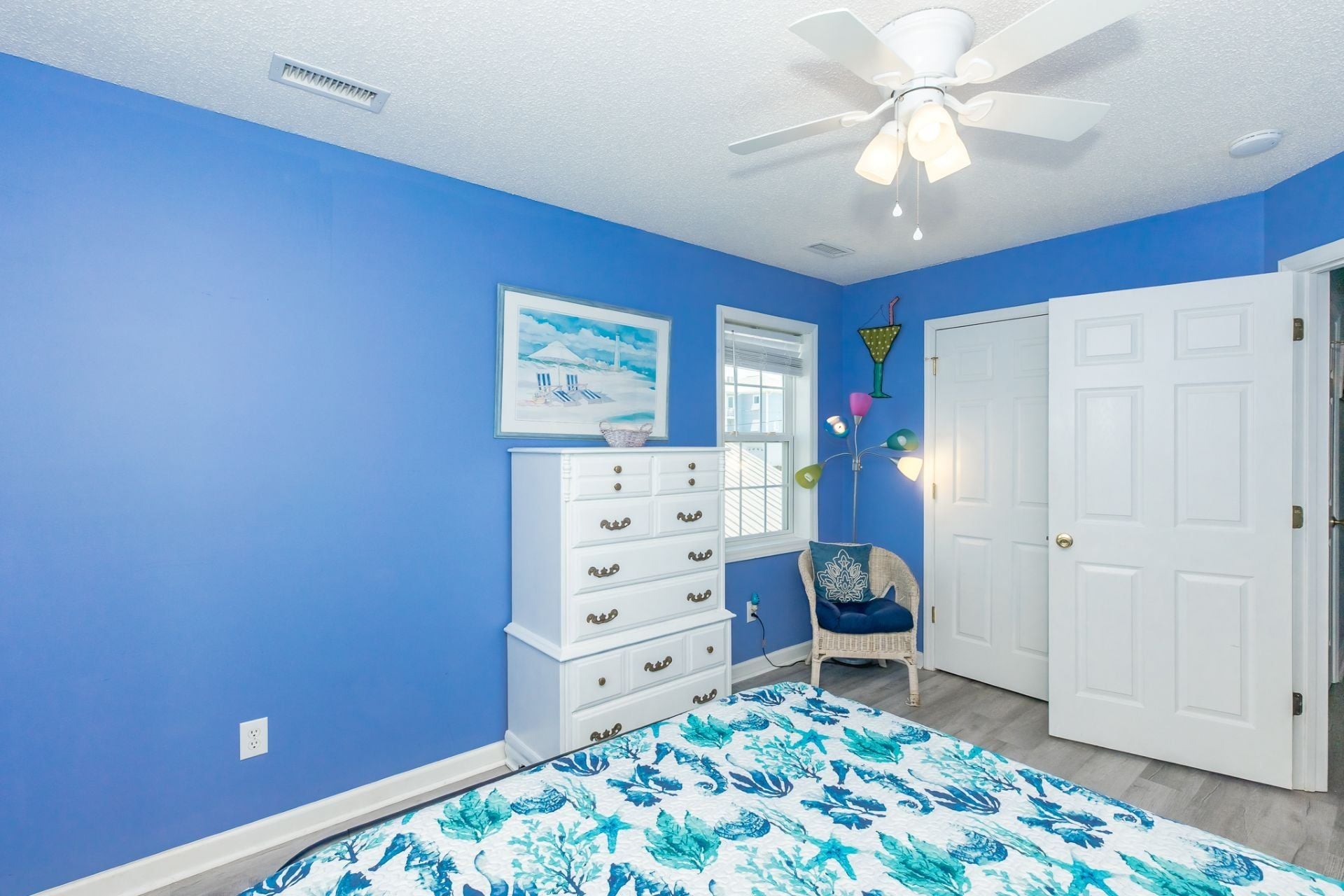 A bedroom with a blue wall, a white dresser, a bed with a floral bedspread, a ceiling fan, and a door.