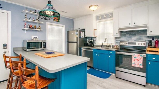 A modern kitchen with a central island, blue cabinets, stainless steel appliances, and wooden chairs. - The Treasure Chest - 3 BR/2 BA Upper Part of Duplex in the Heart of Surf City (Surf City)