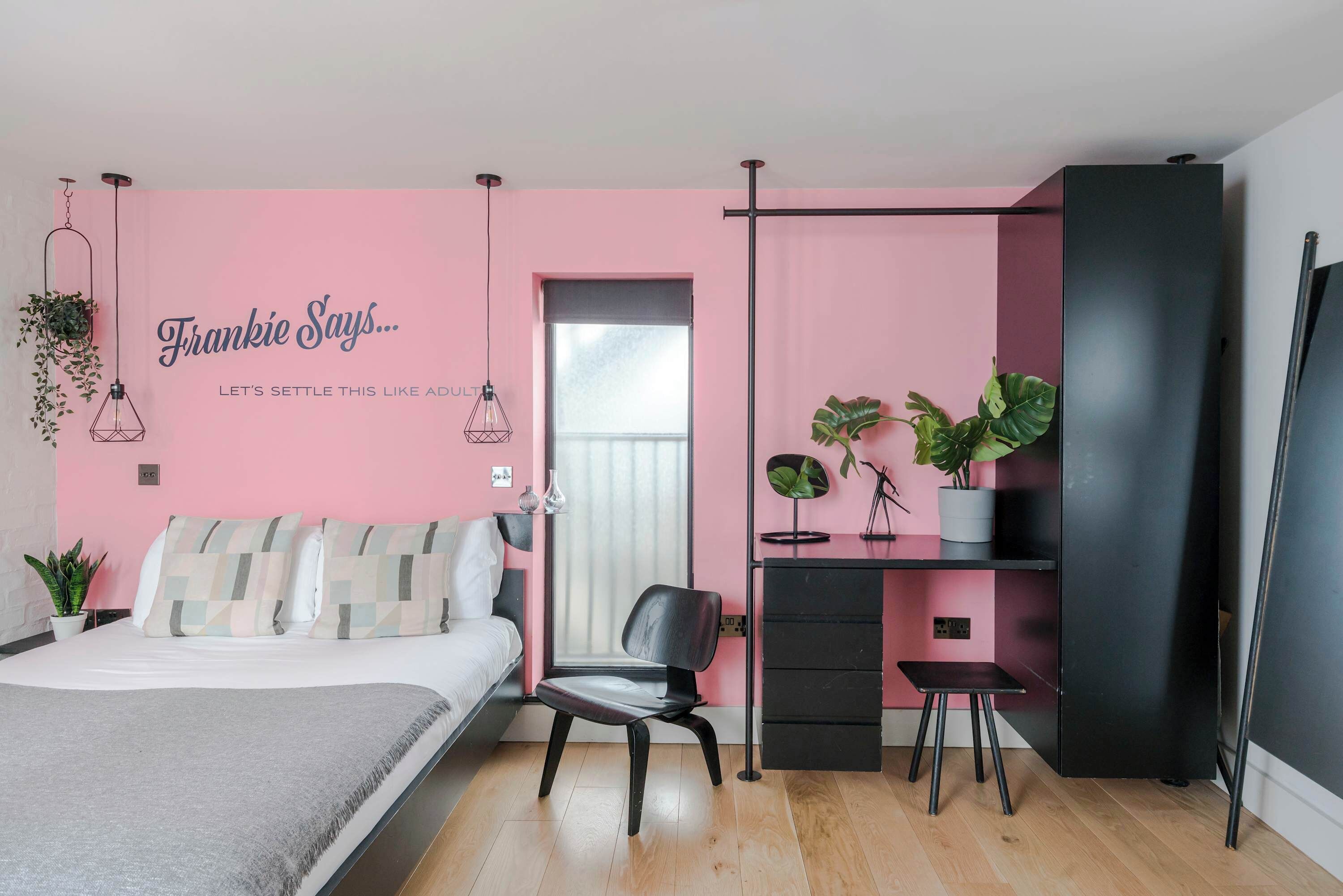Un dormitorio moderno con una pared de acento rosa, una cama con almohadas estampadas, una silla negra, un escritorio con plantas y un armario negro.