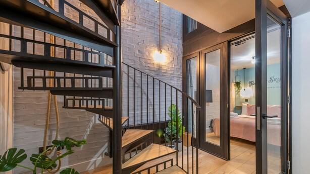 A modern interior with a spiral staircase, a visible bed in the adjacent room, and a brick wall. - Covent Garden 2-Bed Triplex · Sleeps 6 (London)