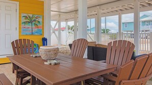 A covered patio with a wooden table and chairs, a painting of a palm tree, and a view of a blue house in the background. - Welcome to the “Pot O’ Gold” at the end of the rainbow! (St. George Island)