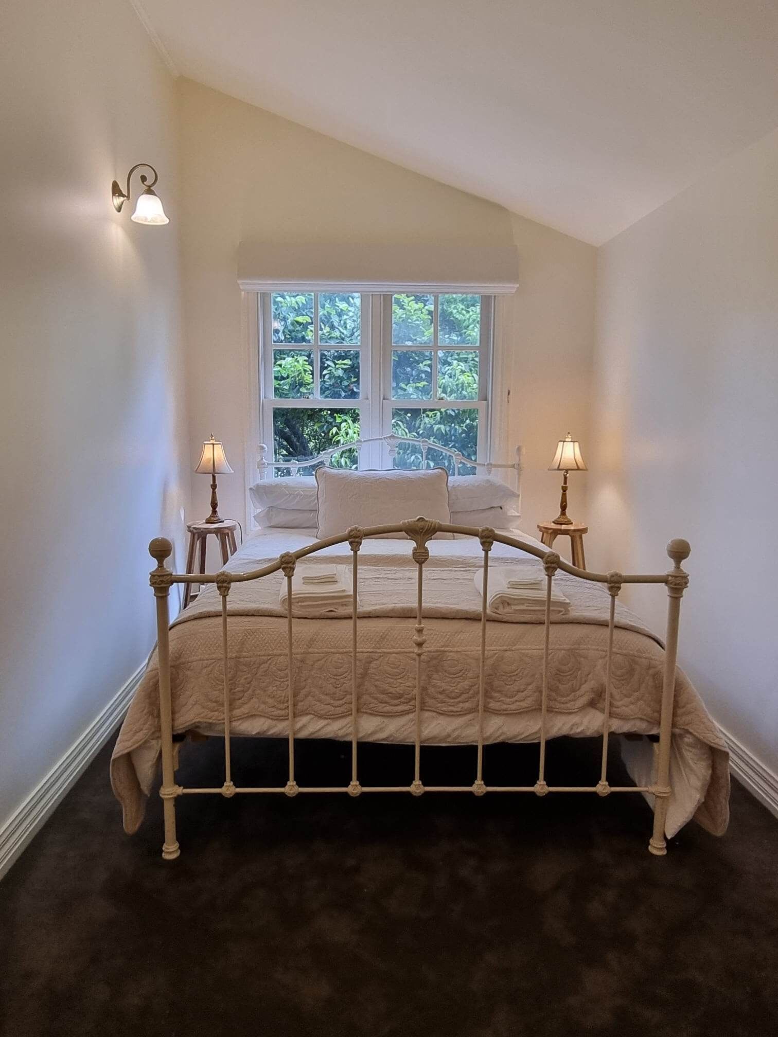 Une chambre à coucher avec un lit, deux lampes de chevet, une fenêtre donnant sur un paysage verdoyant et une petite table.