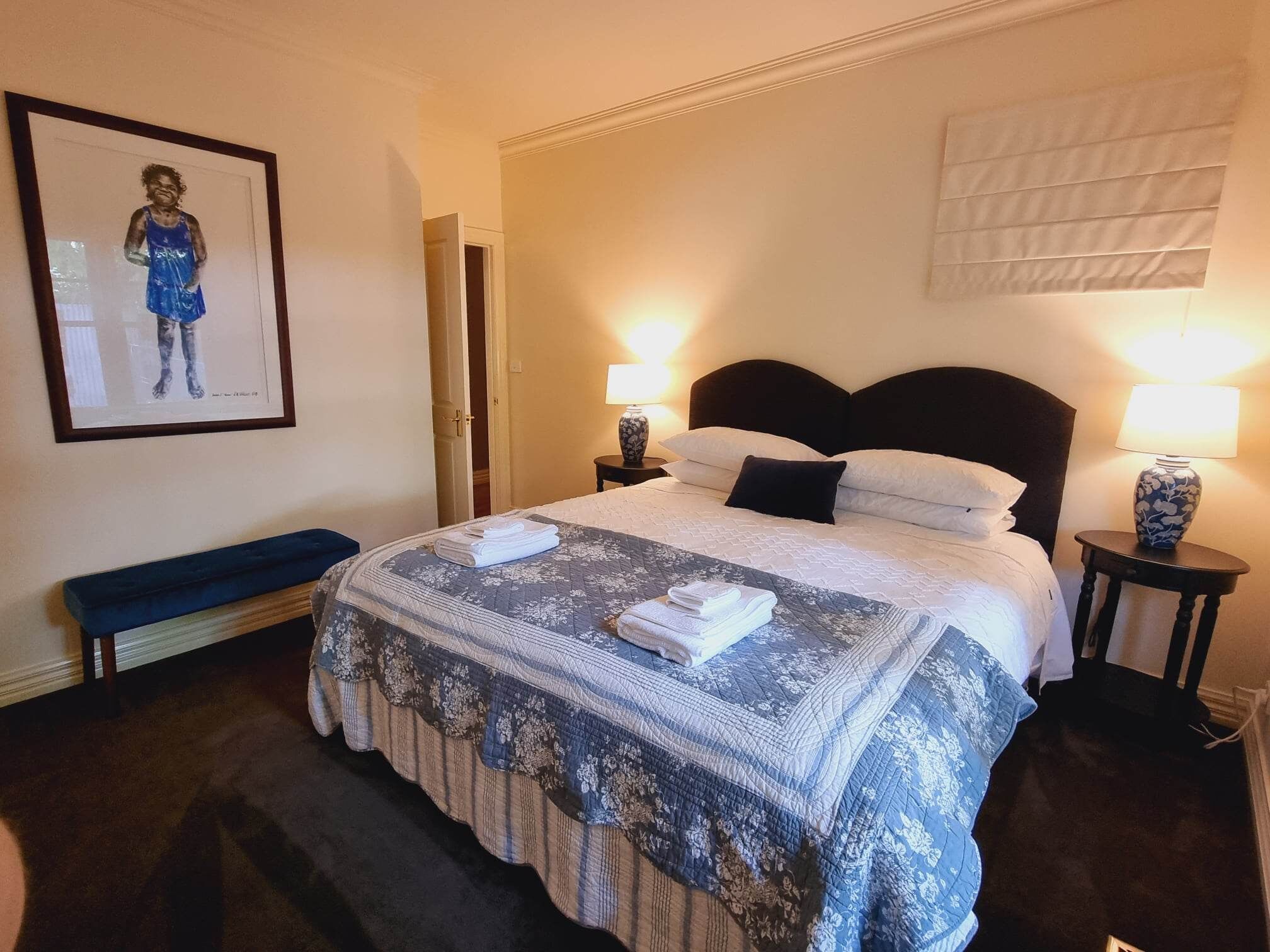 A neatly made bed with a floral-patterned bedspread, two bedside lamps, a framed artwork on the wall, and a bench against the wall.