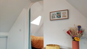 Un dormitori amb sostre inclinat, un llit amb manta taronja, un quadre amb marc a la paret, una cistella teixida i un ram de flors seques en un vas.