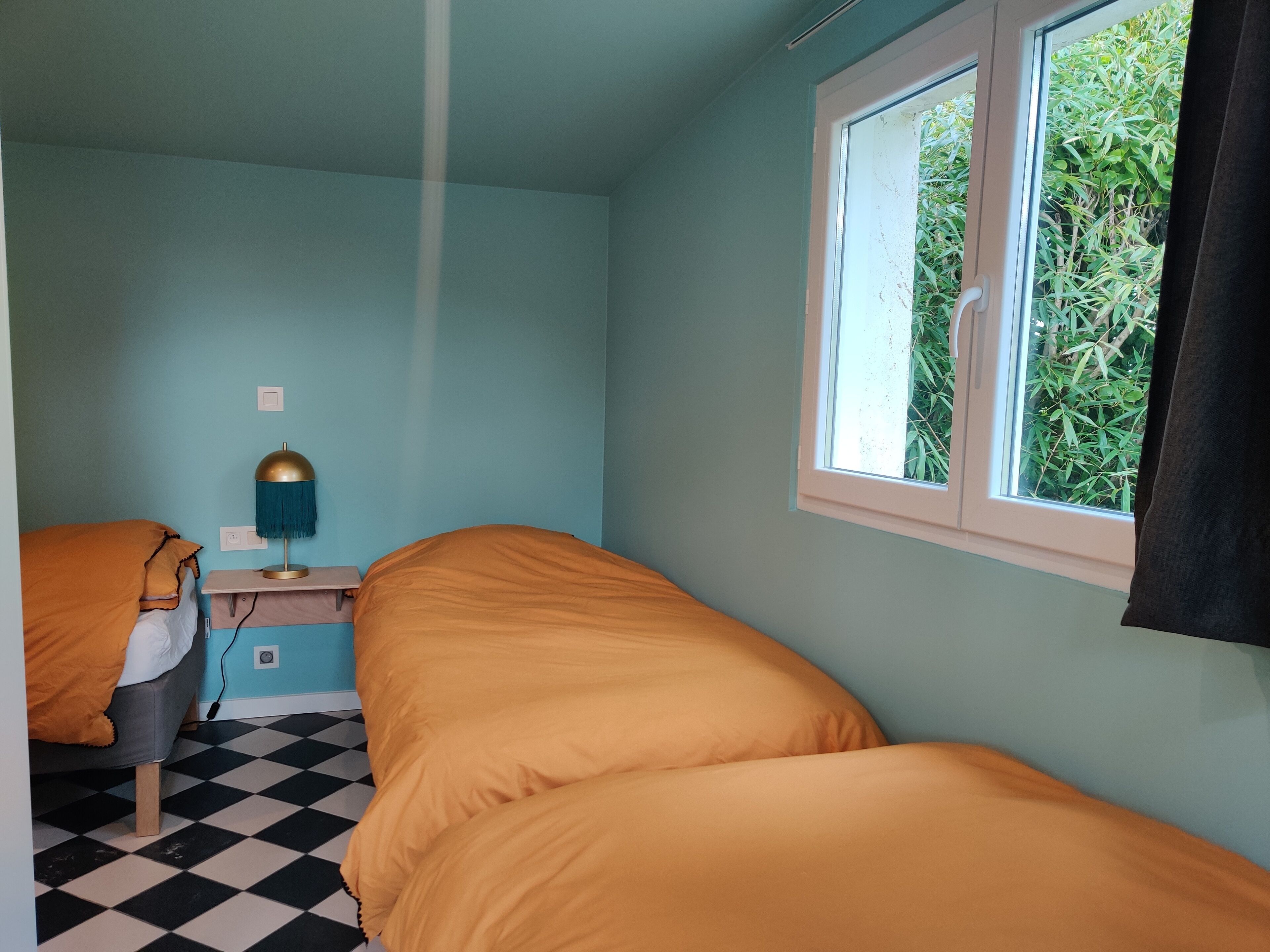 A room with two beds, a window with a view of greenery, a lamp, and a checkered floor.
