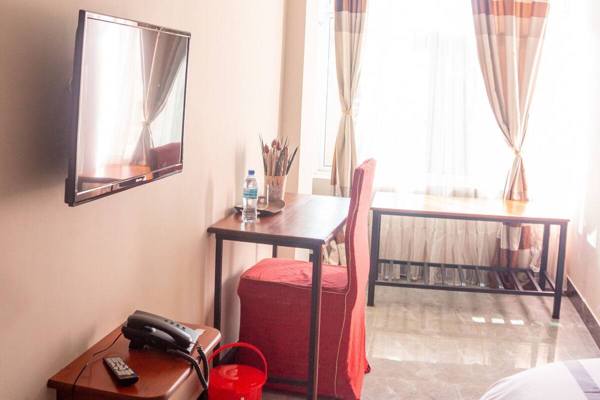 Ett hotellrum med en säng, ett skrivbord med en telefon, en stol, en tv som är monterad på väggen och ett fönster med gardiner.