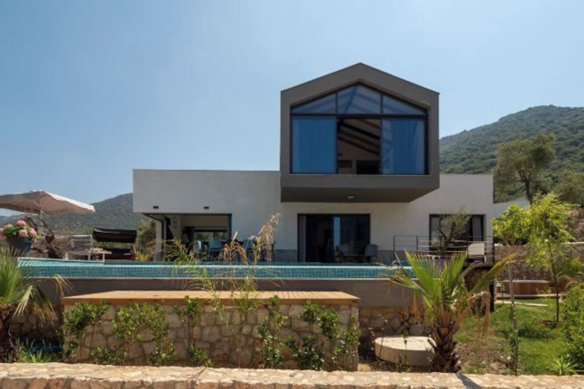 A modern two-story house with a swimming pool, stone walls, and a mountainous backdrop.