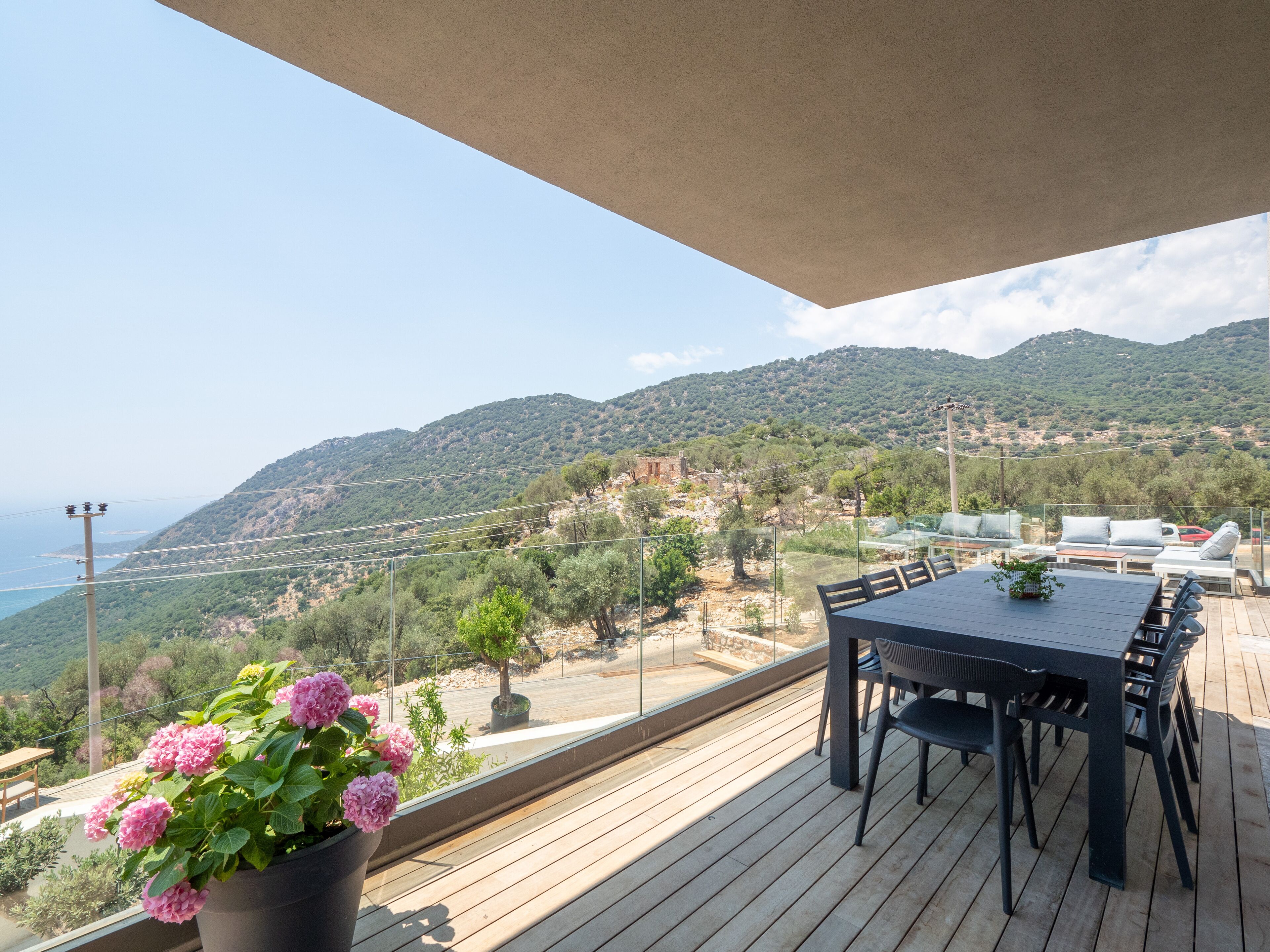 A wooden deck with a table and chairs, overlooking a mountainous landscape and the sea.