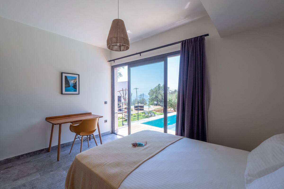 A hotel room with a bed, a desk, a chair, a large window with a view of a pool and outdoor area.