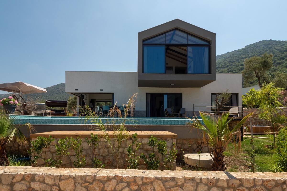 A modern two-story house with a swimming pool, stone walls, and a view of hills.