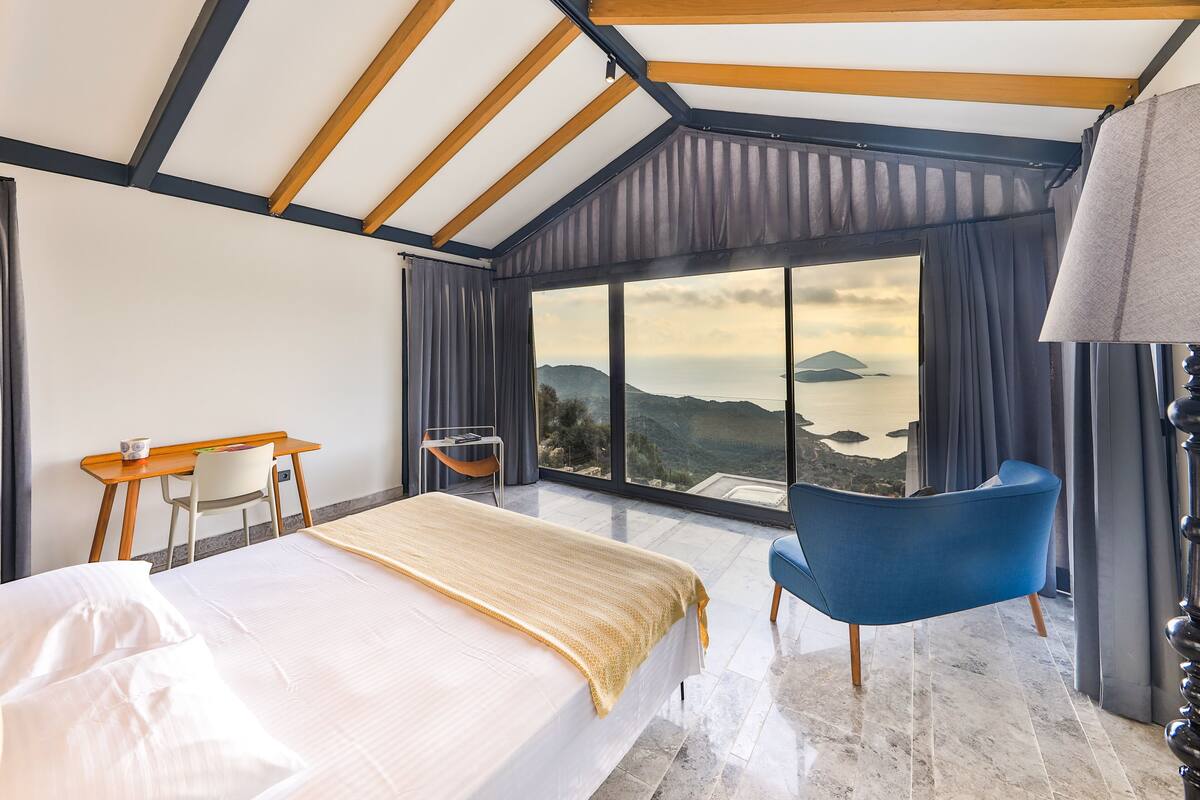 A bedroom with a large bed, a desk, and a chair. There is a view of the outdoors through large windows.