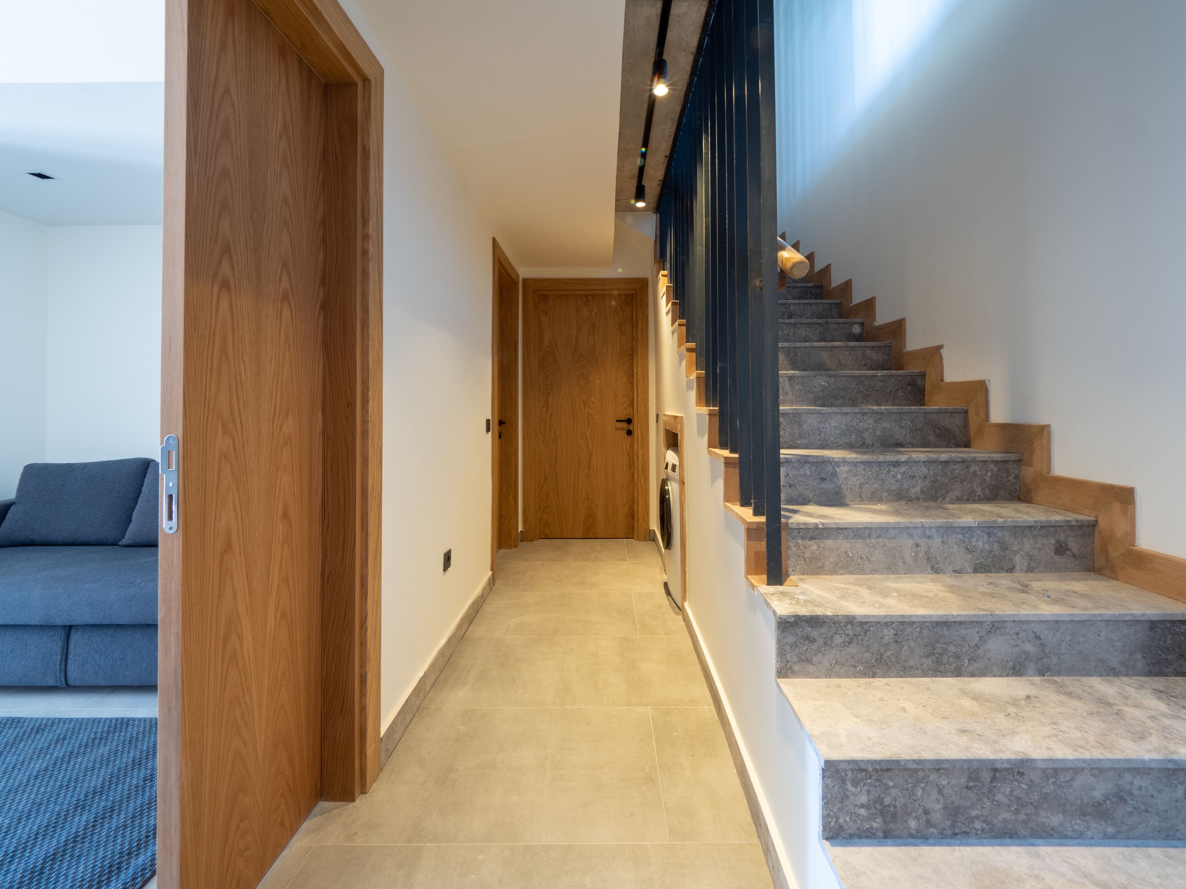 Un intérieur moderne avec une porte en bois, un couloir menant à un escalier avec des marches en pierre et des rampes noires, et un canapé gris en arrière-plan.
