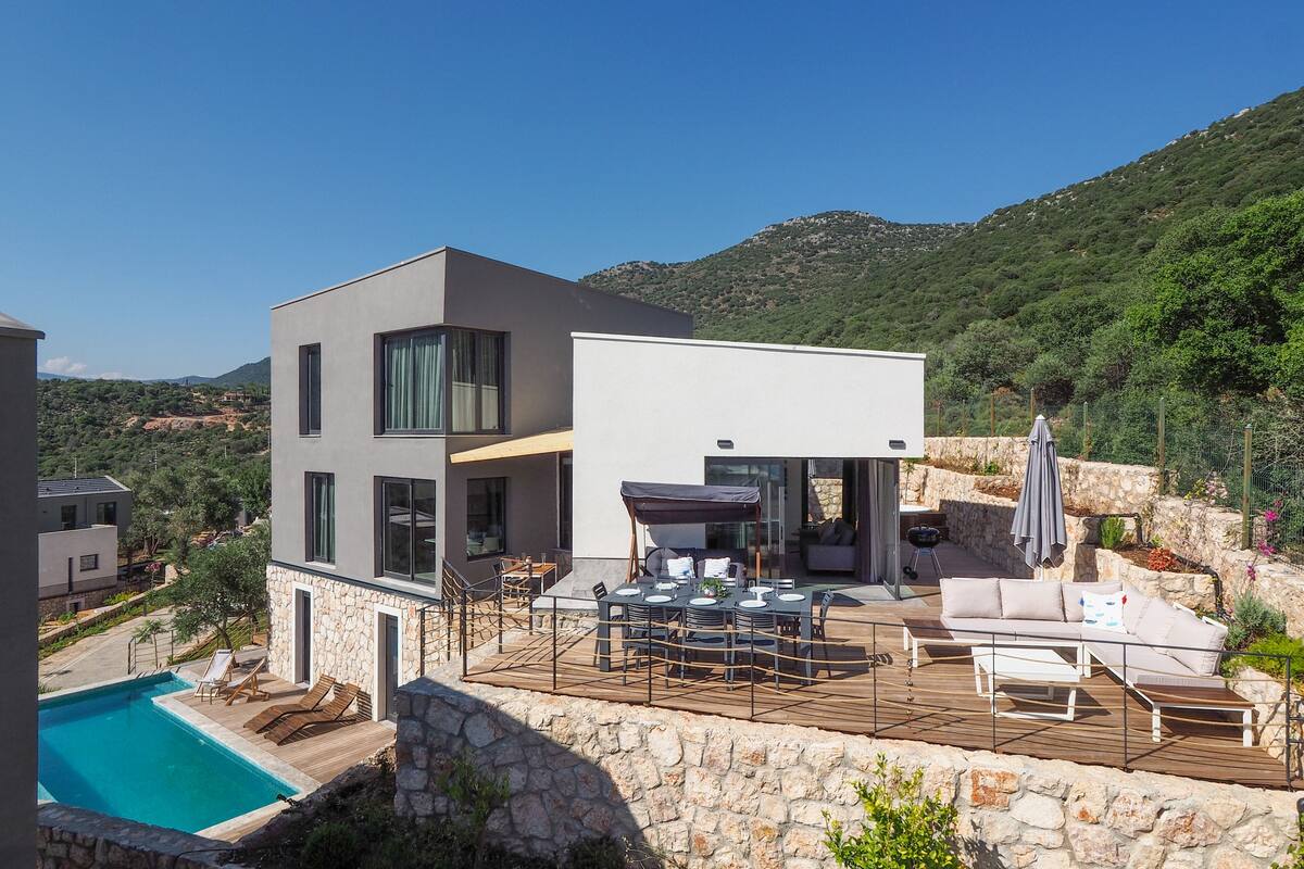 A modern two-story house with a pool, outdoor seating, and a view of the surrounding hills.