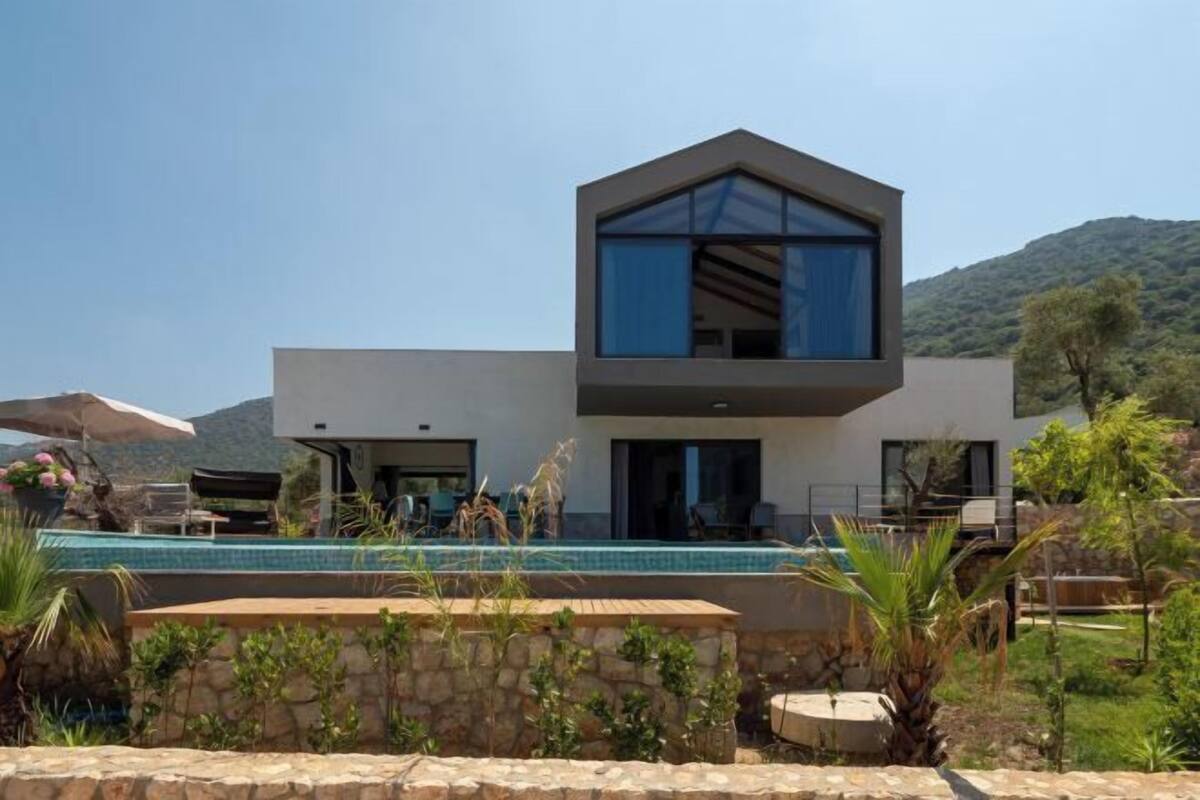 A modern two-story house with a swimming pool, stone walls, and a mountainous backdrop.