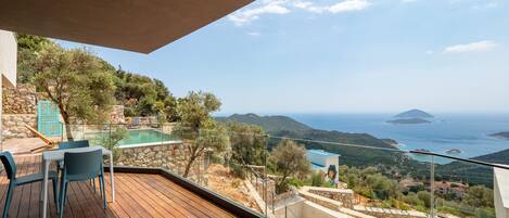A wooden deck with outdoor furniture, a stone wall, and a view of the sea and hills.