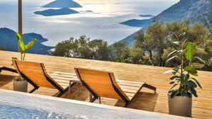 Eine Holzterrasse mit zwei Liegestühlen und einem Blumentopf, mit Blick auf eine Berglandschaft und ein Gewässer.
