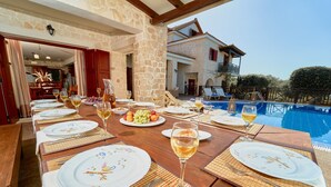 Une table à manger dressée avec des assiettes, des verres et un plateau de fruits, donnant sur une piscine et un bâtiment en pierre.