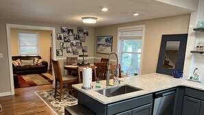 A modern kitchen with a central island, dark cabinetry, and a dining area with wooden chairs and a table. - Cozy Cottage near UNCW, Central Location to All! (Wilmington)