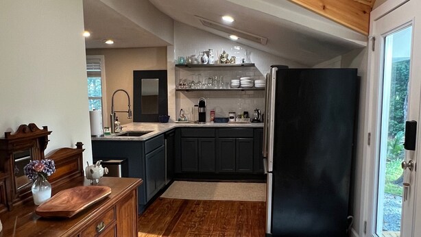 A kitchen with wooden cabinets, a black refrigerator, and a window with a view of greenery. - Cozy Cottage near UNCW, Central Location to All! (Wilmington)