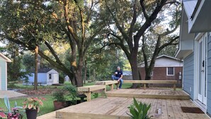 A wooden deck with steps leading to a bench, surrounded by potted plants and trees. - Cozy Cottage near UNCW, Central Location to All! (Wilmington)