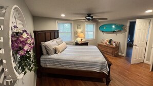 A bedroom with a bed, a ceiling fan, a window with blinds, a nightstand with a lamp, and a surfboard on the wall. - Cozy Cottage near UNCW, Central Location to All! (Wilmington)