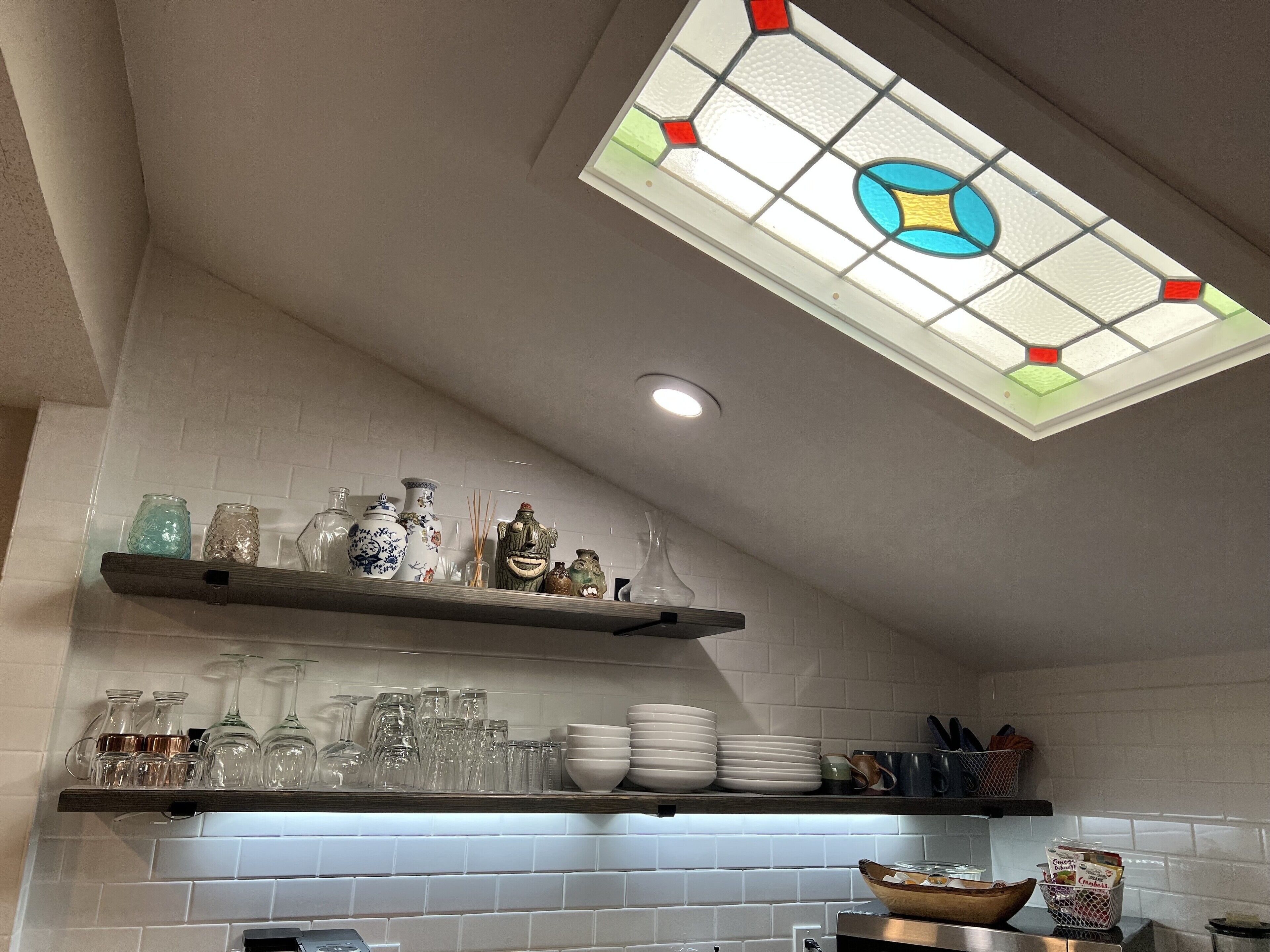 A kitchen with white tiled walls, a stained glass window, and shelves holding glassware and dishes.