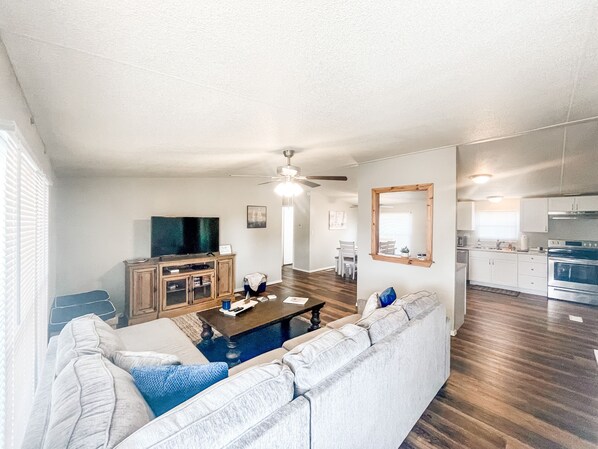 A spacious living room with a large sofa, a coffee table, and a TV. There is a dining area with a table and chairs. The kitchen is visible with cabinets and appliances.