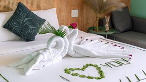 Cama bem arrumada com toalhas dispostas em formato de cisne, uma decoração com folha em forma de coração e uma pequena mesa com um vaso de flores.