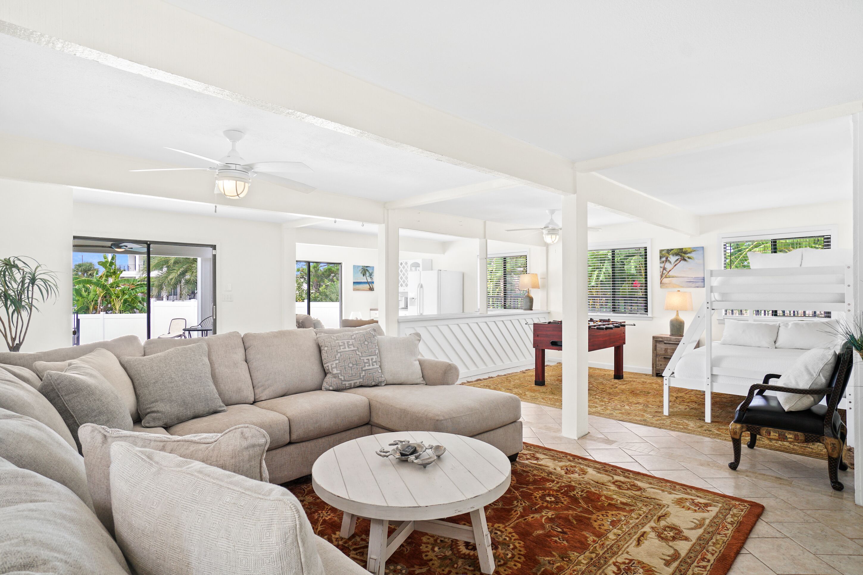 A spacious living area with a beige sectional sofa, a round coffee table, and a patterned rug. There is a wooden chair and a white bench in the background.