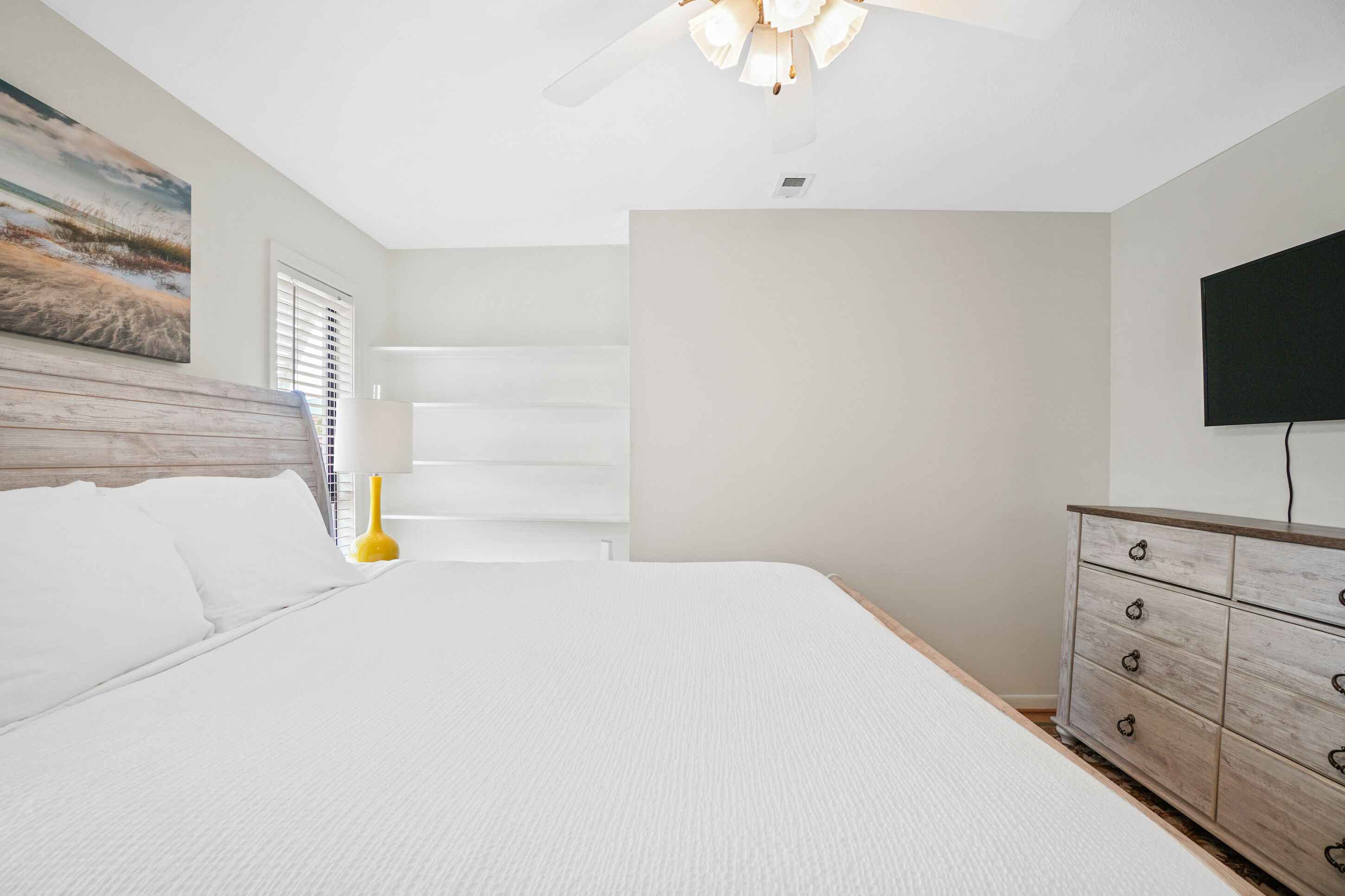 A bedroom with a large bed, a wooden headboard, a bedside table with a lamp, a dresser, and a painting on the wall.