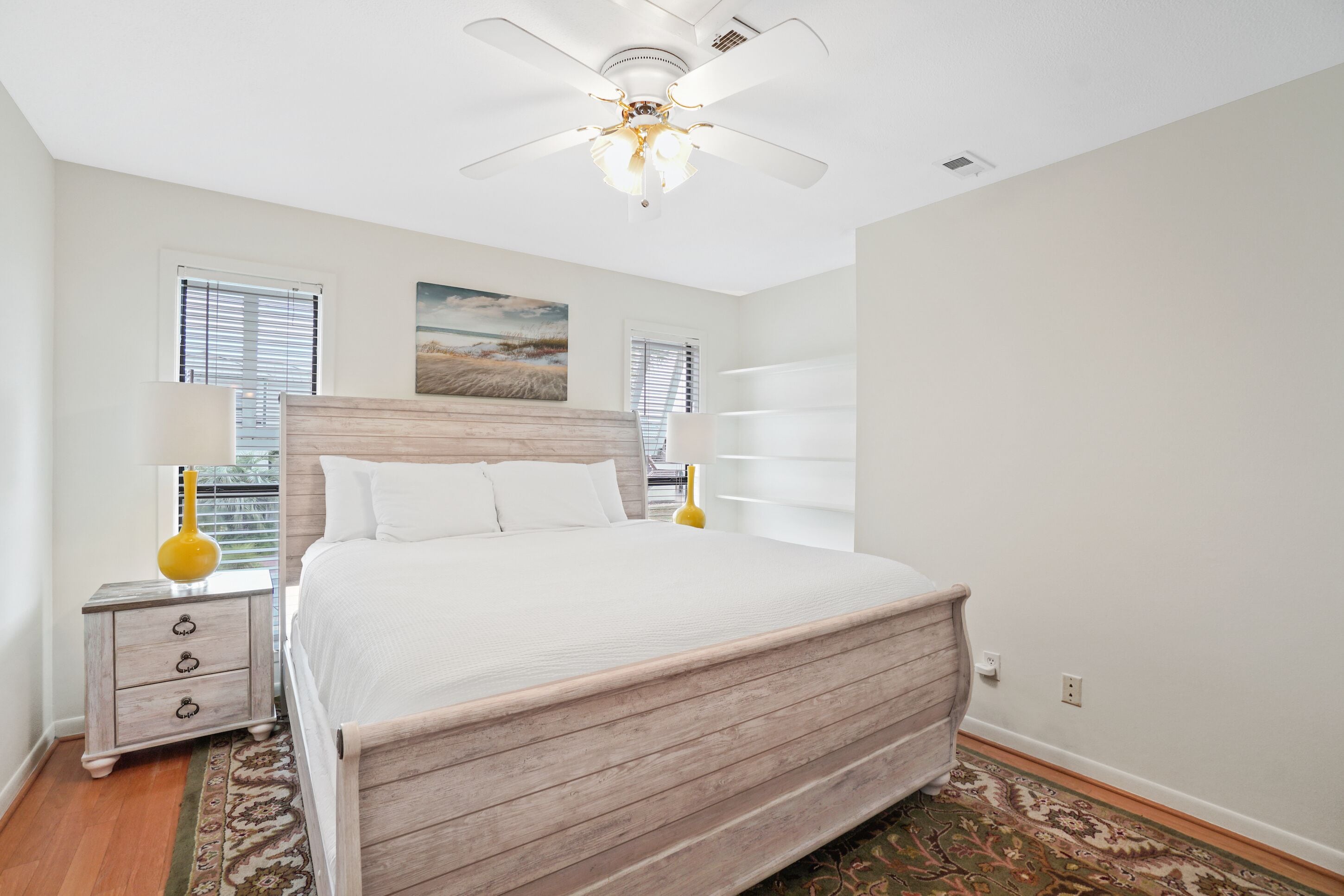 A bedroom with a wooden bed, white walls, a ceiling fan, and a nightstand with a lamp.
