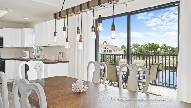 A modern kitchen with white cabinets, a wooden dining table, and a view of a balcony with outdoor seating.