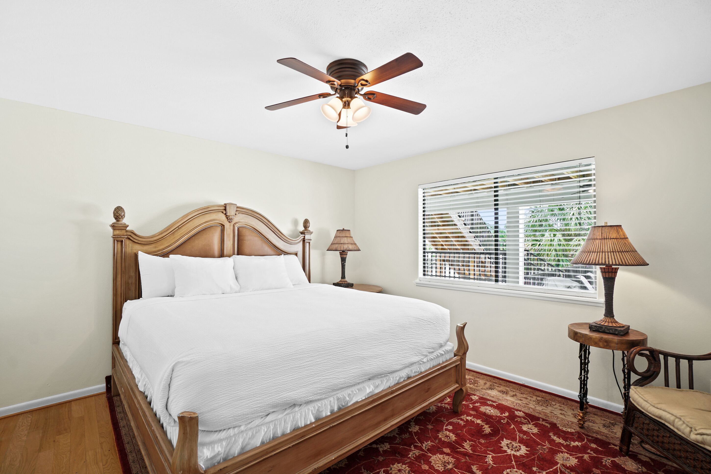 A bedroom with a wooden bed, a ceiling fan, a window with blinds, two bedside lamps, and a red patterned rug.