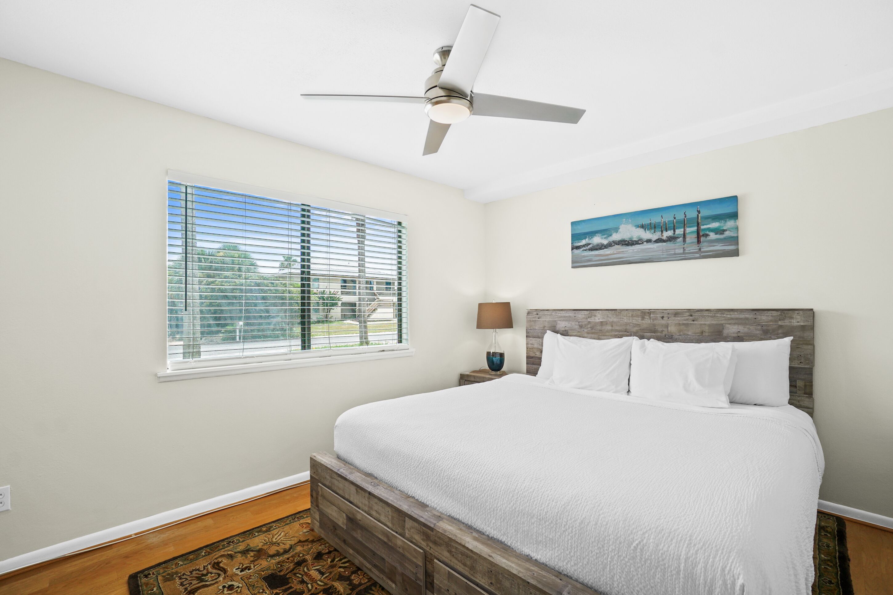 A bedroom with a bed, a ceiling fan, a window with blinds, a painting on the wall, and a nightstand with a lamp.