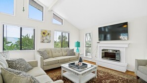A living room with a beige sofa, a coffee table, a flat-screen TV mounted above a fireplace, and large windows with blinds.