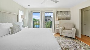 A hotel room with a large bed, a ceiling fan, a view of a pool and greenery, a chair, and a small table with a lamp.