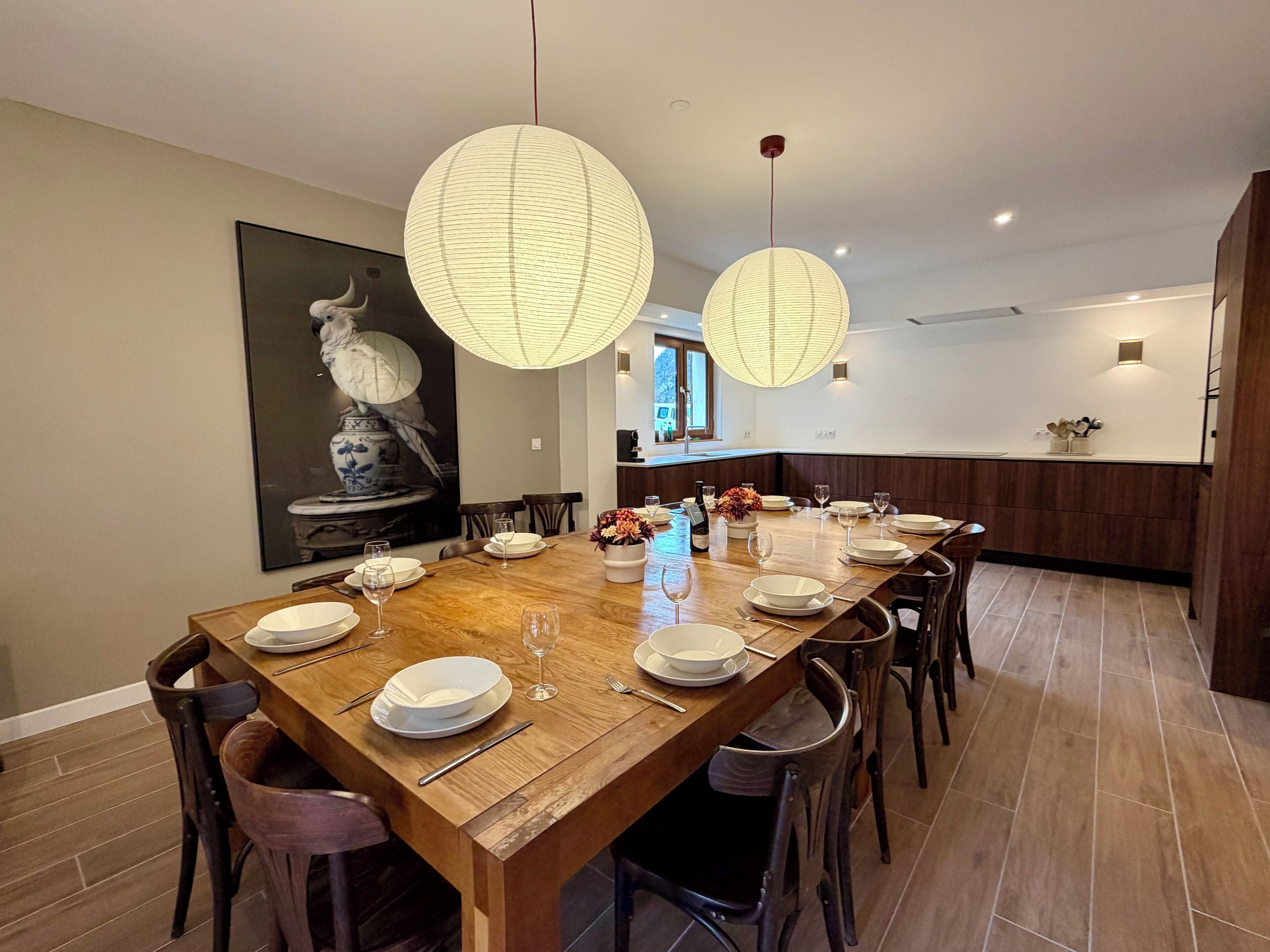 Un'area pranzo con un tavolo di legno apparecchiato per sei persone, circondato da sedie. Si notano lanterne di carta appese, un quadro al muro e una zona cucina sullo sfondo.