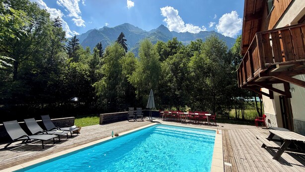 A swimming pool with lounge chairs and tables, surrounded by trees and mountains in the background. - Chalet les Ecrins - 230m2 ✨ Sauna - Swimmingpool - Garden ✨  (Vénosc, Les Deux Alpes)