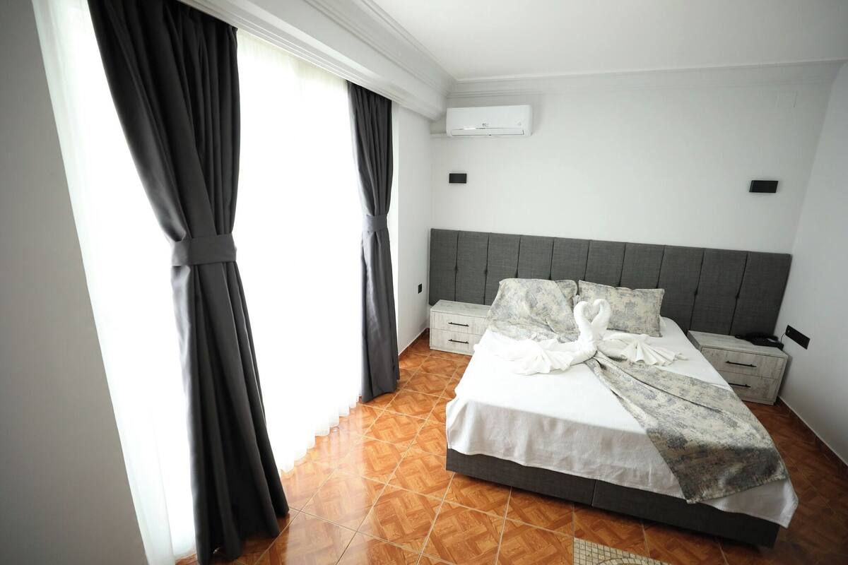 A hotel room with a bed, a nightstand, a wall-mounted air conditioner, and a window with curtains.