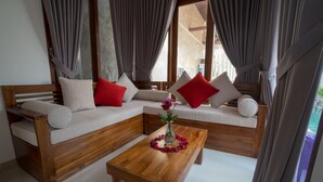 A modern living room with a wooden sofa, red and white cushions, a wooden coffee table with a vase of flowers, and large windows with curtains. - Royani 1-Bedroom Private Pool Villa (Gianyar)